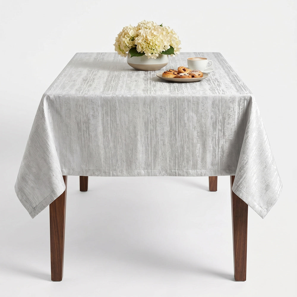 Silver Tablecloth featuring a textured design, elegantly draped over a wooden table with flowers and snacks.