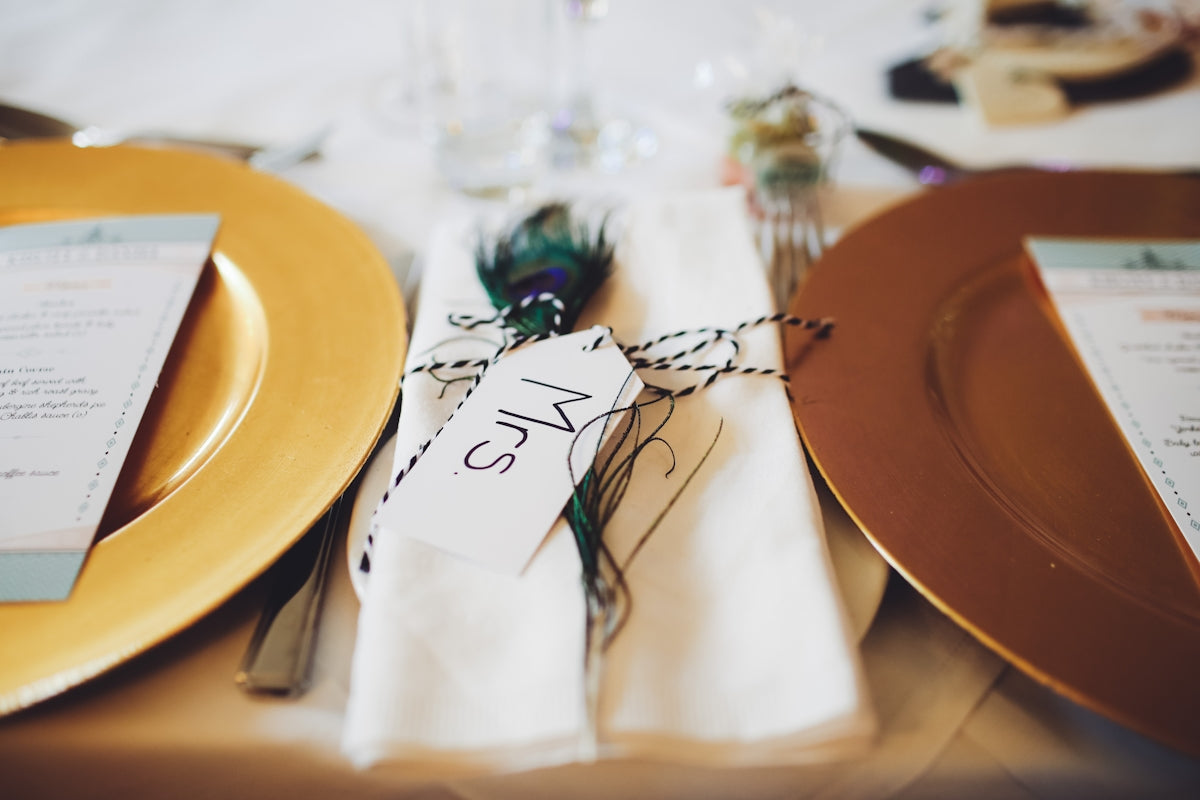 Elegant gold charger plates with menu cards and decorative peacock feathers for a wedding table setting.