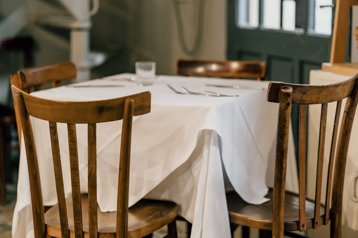 Wooden dining table with white tablecloth and chairs in a cozy restaurant setting.