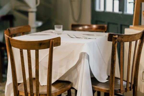 Wooden dining table with white tablecloth and chairs in a cozy restaurant setting.
