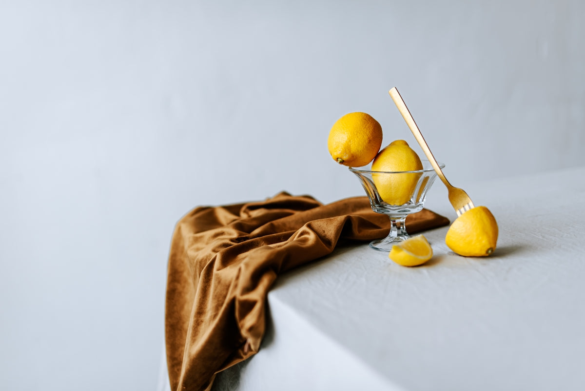 Fresh lemons in a glass bowl with a wooden spoon on a textured tablecloth.