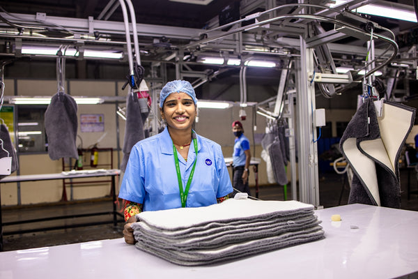 Product name not provided. Smiling worker in a blue uniform with a hairnet, standing behind a stack of clean linens.