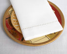 Folded white dinner napkin with a drawn-thread hemstitch border on a decorative gold plate