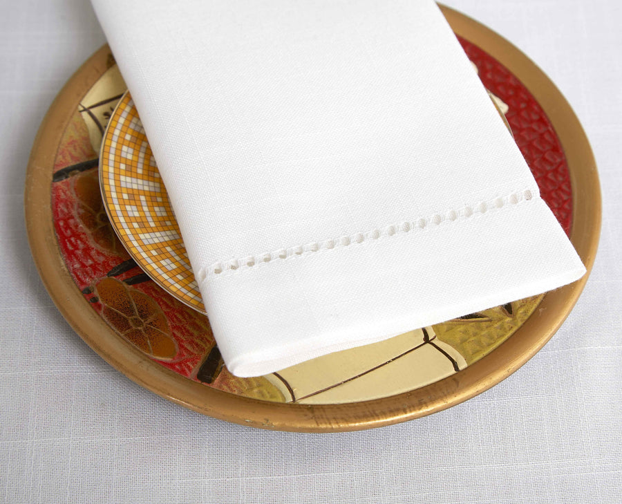 Folded white dinner napkin with a drawn-thread hemstitch border on a decorative gold plate